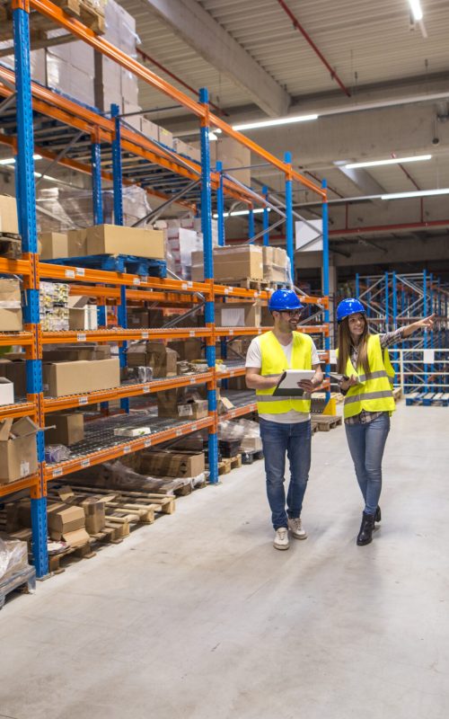 Two caucasian warehouse workers walking in distribution storage area discussing about logistics and organization.